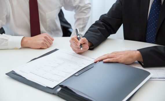 A Person in Black Suit Holding a Pen Near the Documents on the Table